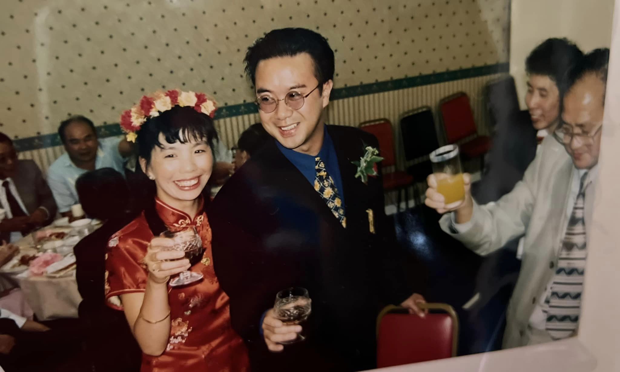Angela and Gordon on their wedding, glasses in hand making a toast by a dining table of guests